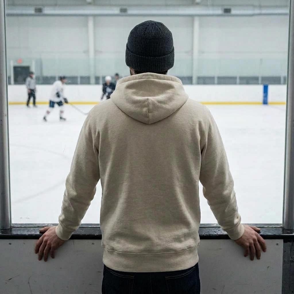 Rear view of a man in a plain heavyweight hoodie and knit toque at the rink boards, looking out over the ice